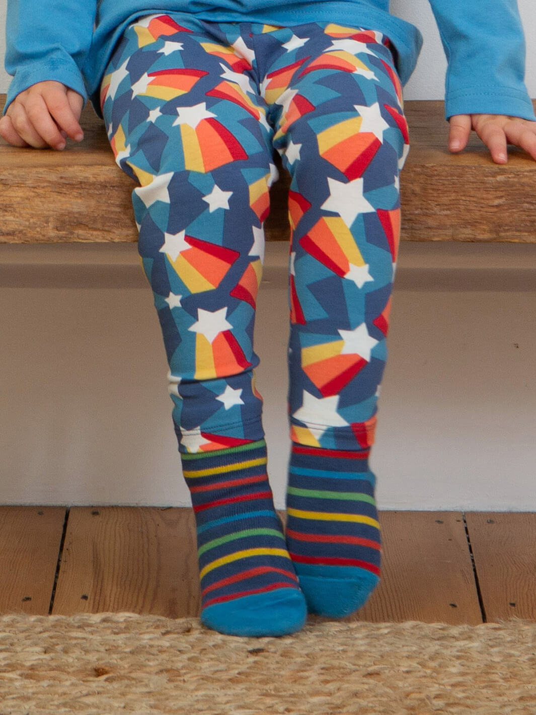 Niño sentado usando leggings de estrellas con calcetines de rayas azules y rojas antideslizantes de algodón orgánico.