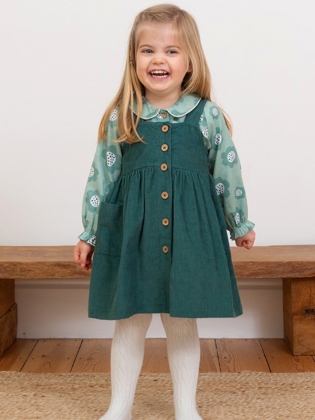 Niña vistiendo jardinera de algodón orgánico verde pino, combinada con una blusa verde y pantys blancas, logrando un look cómodo y elegante.