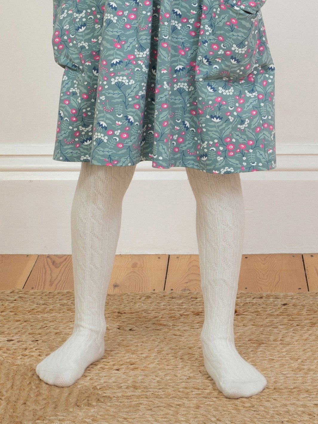 Niña usando pantys de algodón orgánico en color crema con diseño de punto trenzado, combinadas con un vestido para un look elegante y acogedor.