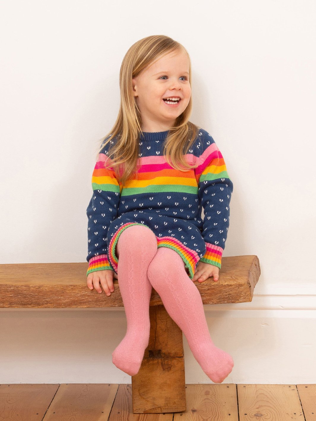 Niña sonriendo mientras usa el vestido tejido azul marino con detalles de arcoíris, mostrando su ajuste cómodo y estilo versátil.