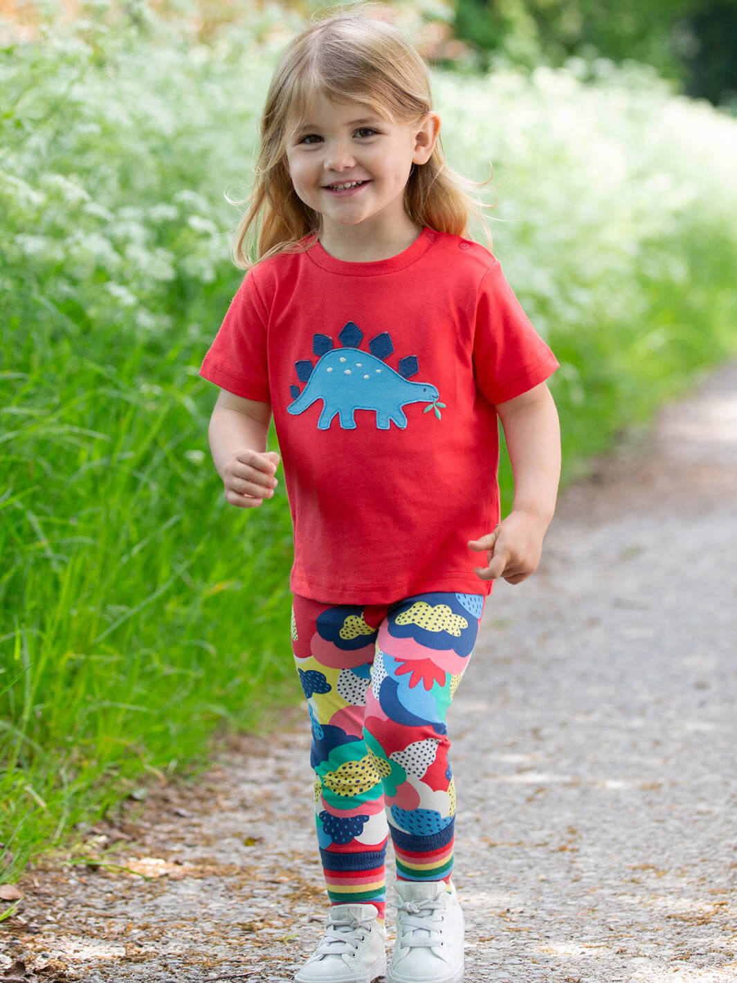 Niña usando polera roja de algodón orgánico con dinosaurio azul, combinada con leggings de colores.